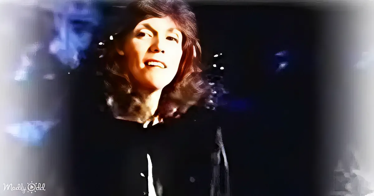 A person with long, dark hair and a black hoodie is illuminated by a spotlight against a dark background.