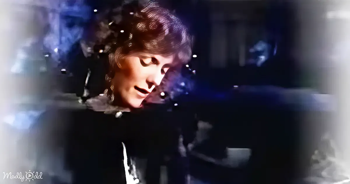 A person with short hair and a dark outfit is seen in a dimly lit environment, possibly a performance or event.