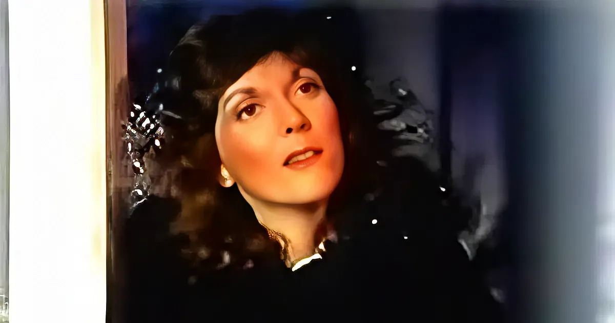 A woman with dark, curly hair and a concerned expression, wearing a dark jacket, stands in front of a broken window.