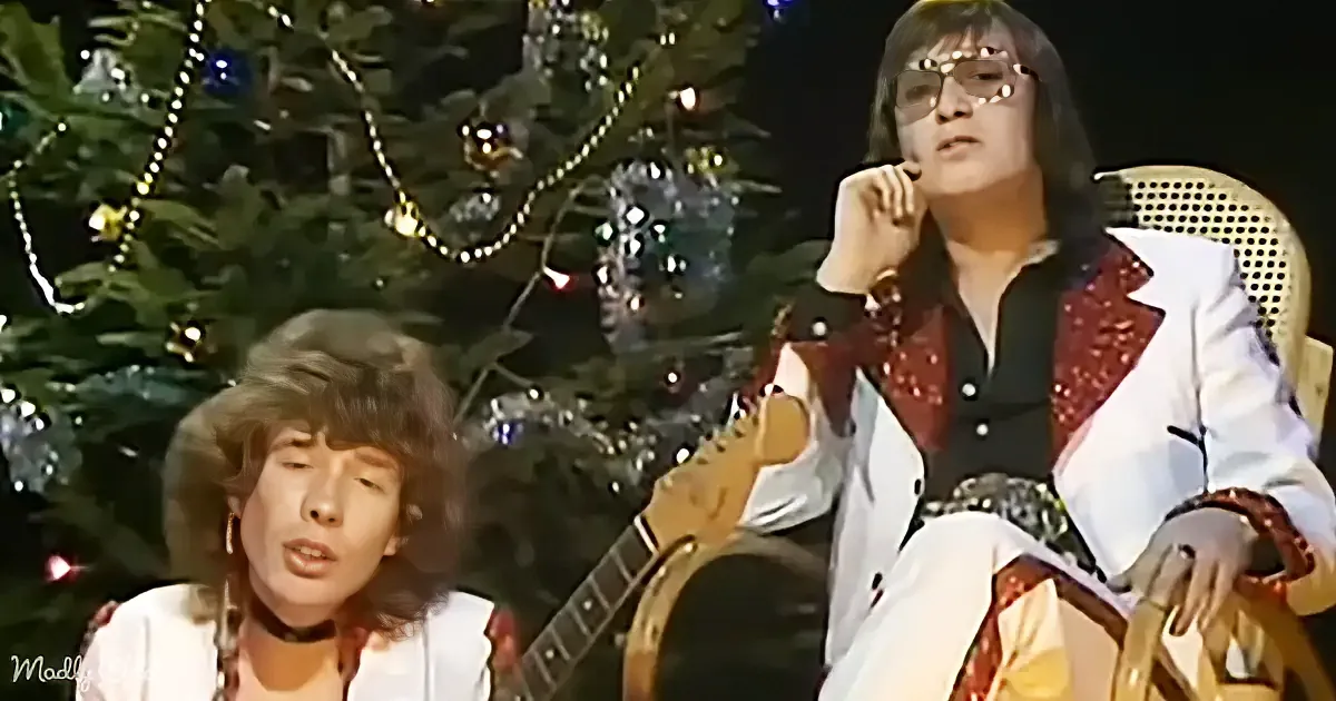 A man in a white suit with red accents sits next to another man playing a guitar in front of a decorated Christmas tree.