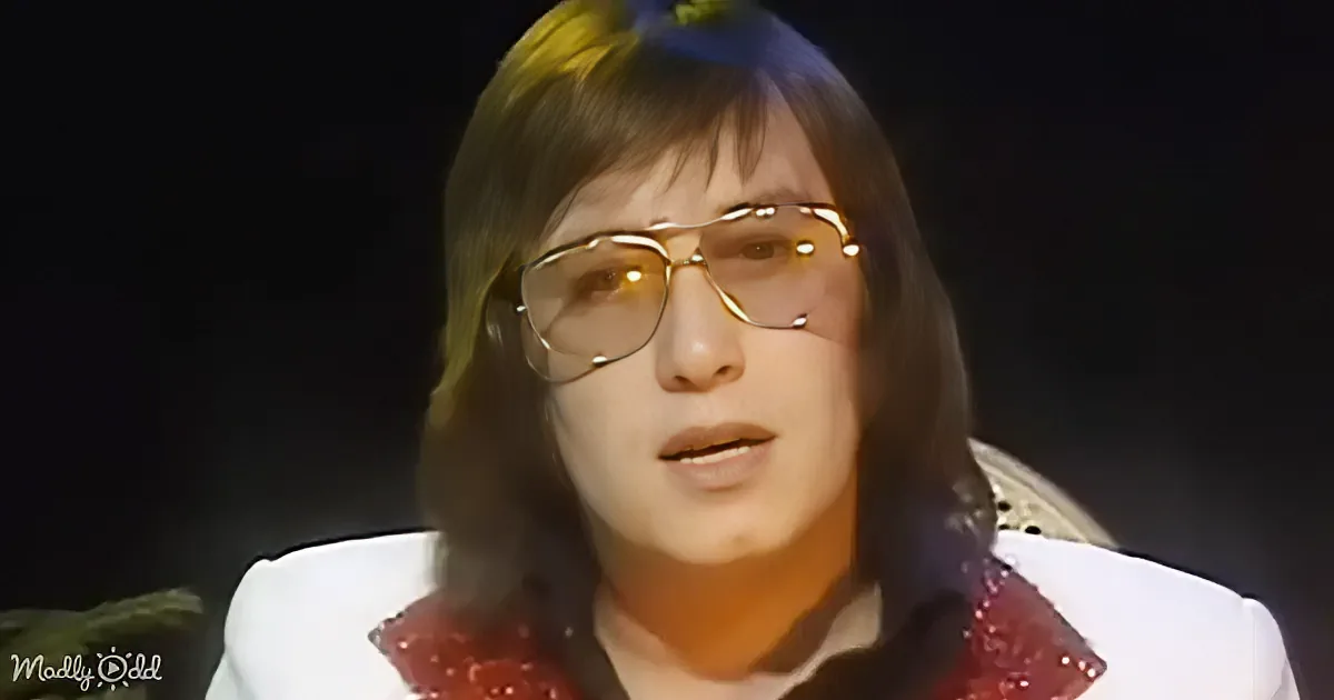 A person with long hair and glasses, wearing a red and white outfit, is speaking into a microphone.