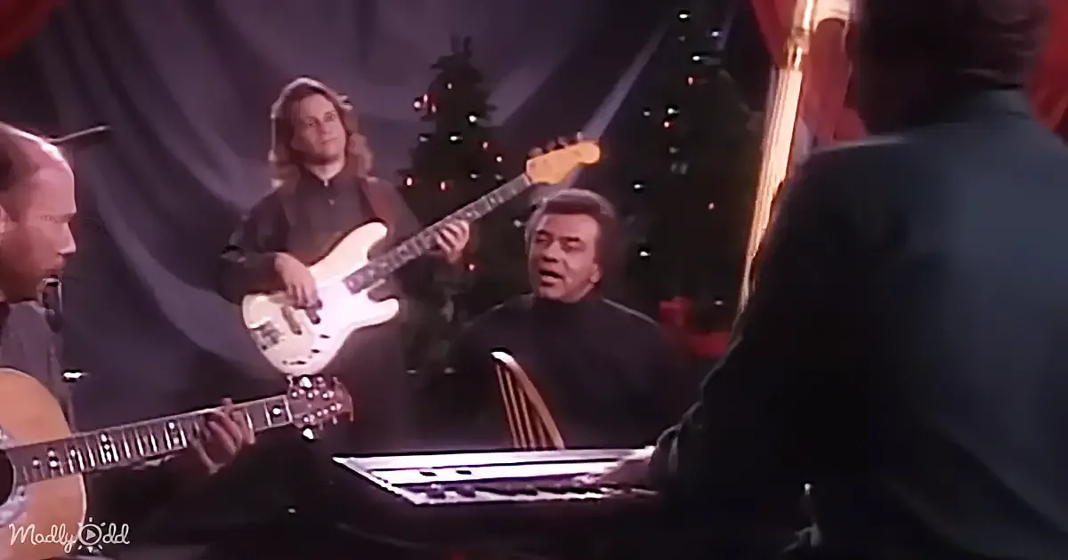 A musician plays guitar while another sings in front of a Christmas tree, with a keyboardist and guitarist in the foreground.