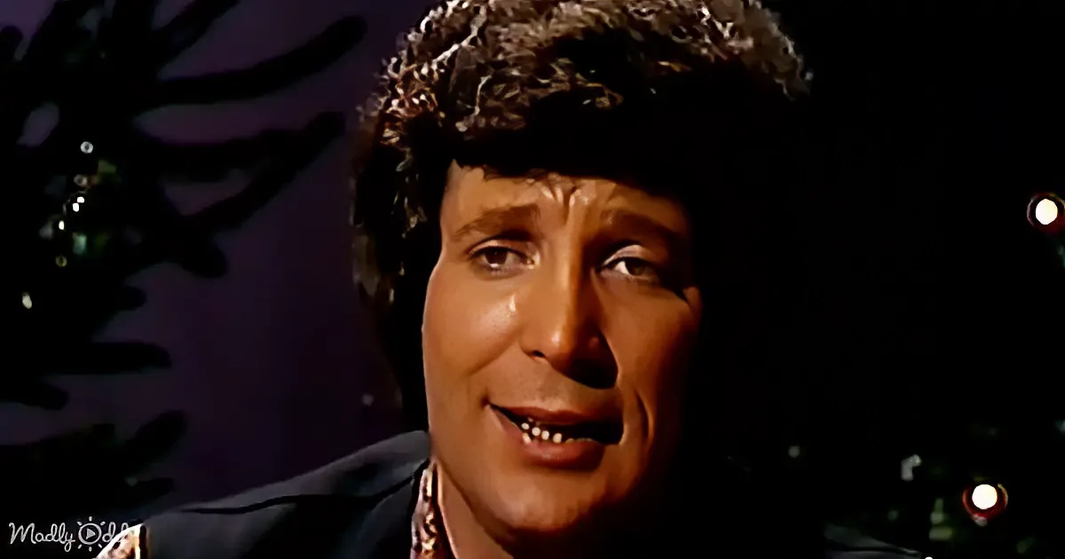 A person with curly hair and a dark outfit is smiling, with a dark background and some blurred lights.