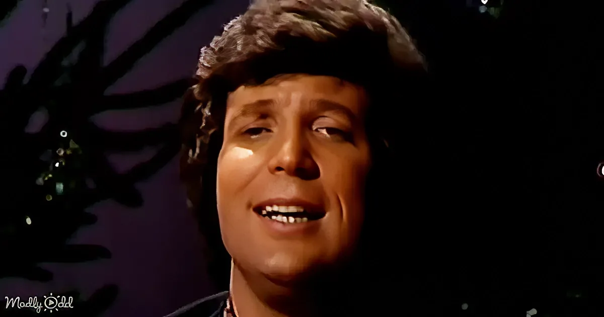 A person with curly hair and a dark background, possibly singing or speaking.