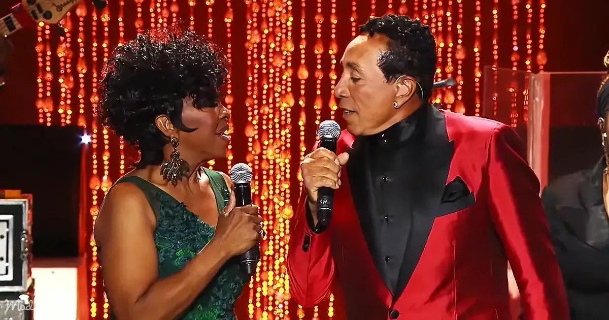 A man in a red suit and a woman in a green dress sing into microphones on a stage with glowing lights.