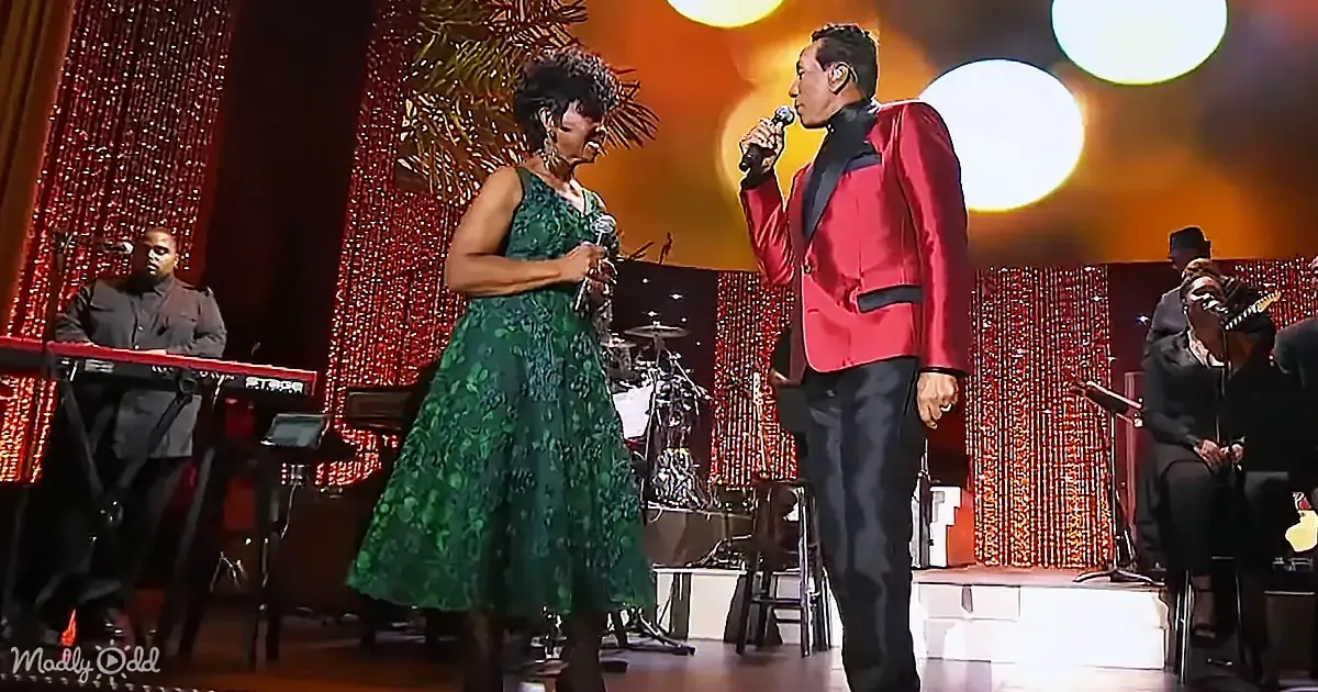 A woman in a green dress and a man in a red suit sing on stage with a band in the background.