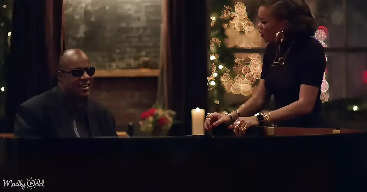 A man in sunglasses sits at a piano, while a woman in black gestures beside him, with a lit candle and Christmas tree in...