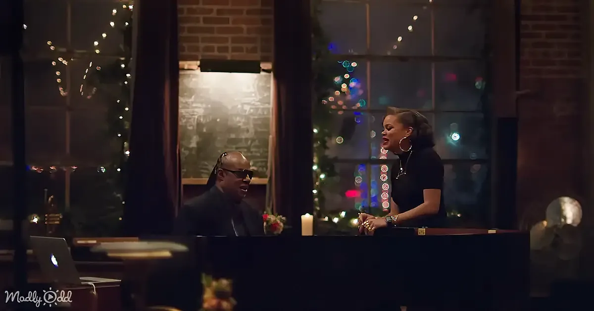 A man and woman are seated at a piano, with Christmas lights and a chalkboard in the background.