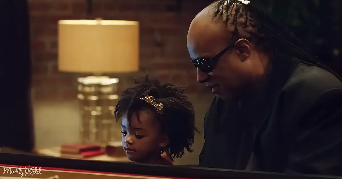 A man and a young child are sitting at a piano, with the man wearing sunglasses and the child looking down.