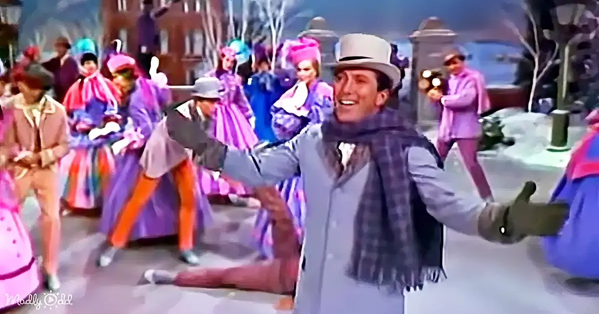 A man in a gray suit and scarf dances joyfully in a colorful, festive setting with other people in vibrant costumes.