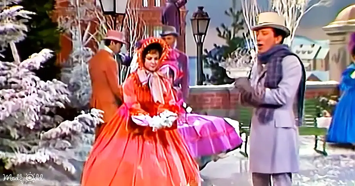 A woman in a vibrant red Victorian dress stands with a man in a gray coat, surrounded by snowy trees and other people in...