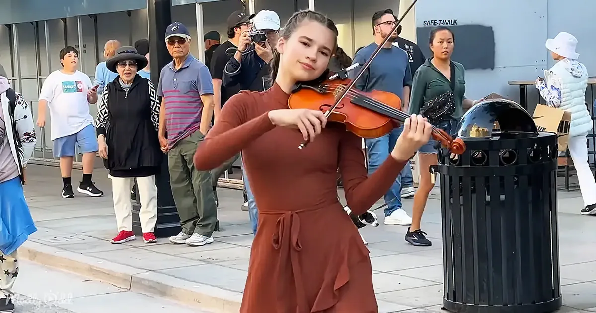 A young woman in a brown dress plays the violin on a sidewalk, surrounded by onlookers.
