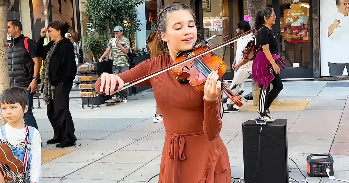 A young woman in an orange dress plays the violin on a busy sidewalk, surrounded by passersby.
