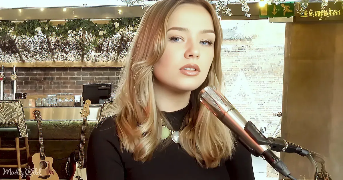 A woman with blonde hair sings into a microphone in a cozy, decorated room with guitars and a brick wall.