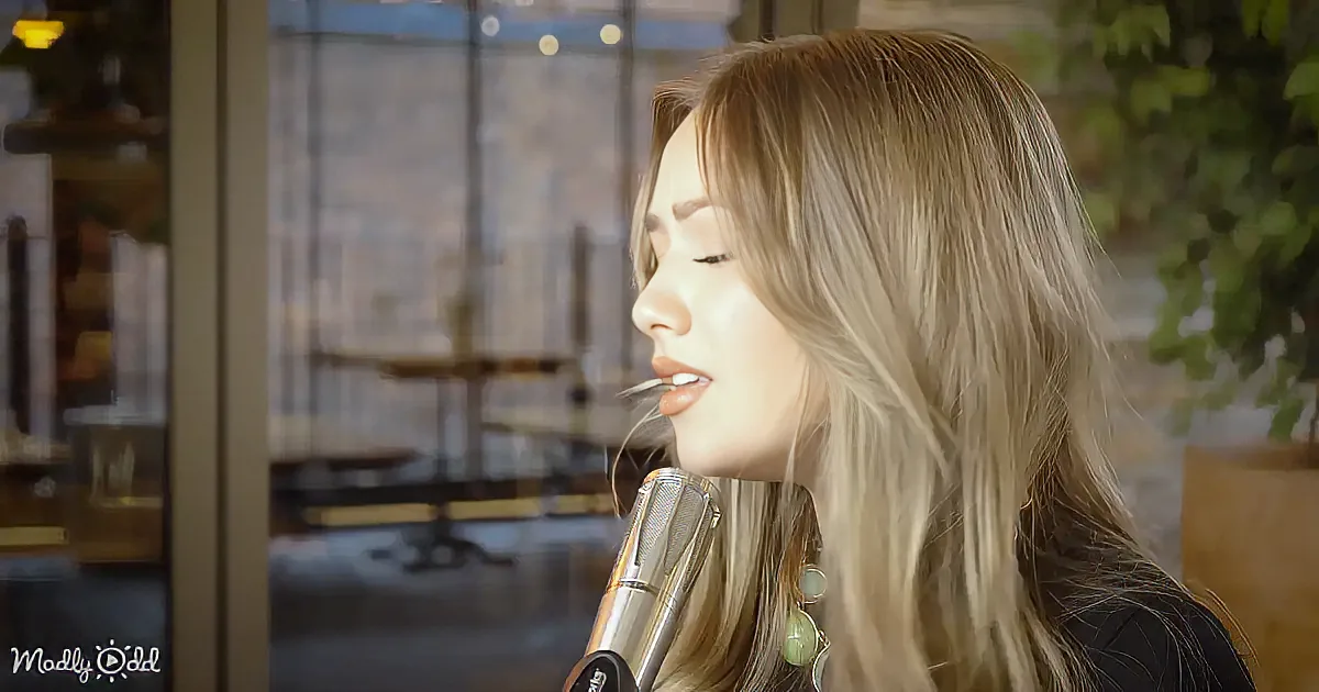 A woman with long blonde hair sings into a microphone in a softly lit indoor setting.