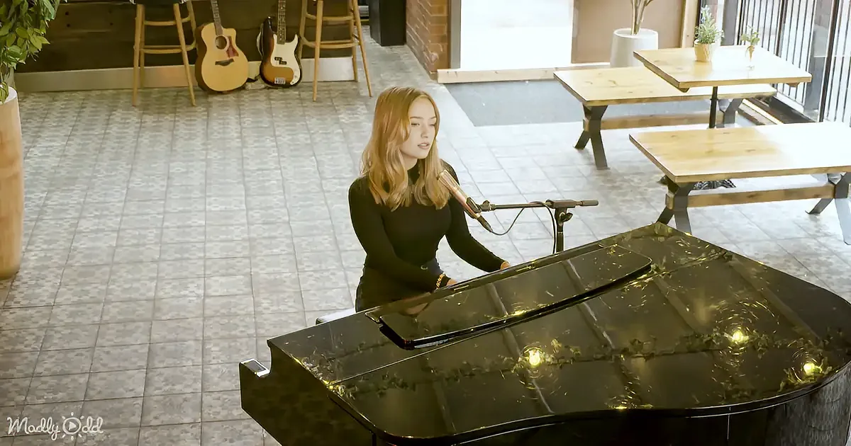 A woman sings into a microphone while playing a grand piano in a cozy room with guitars and tables.
