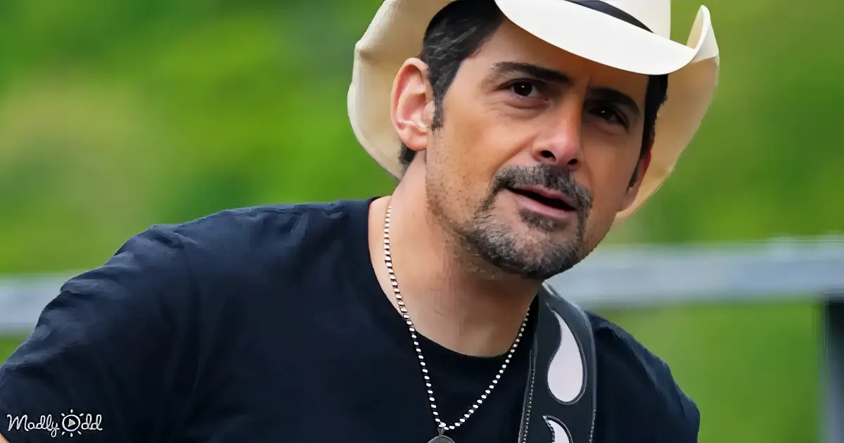A man in a cowboy hat plays an acoustic guitar on a bridge with a scenic forest and river backdrop.