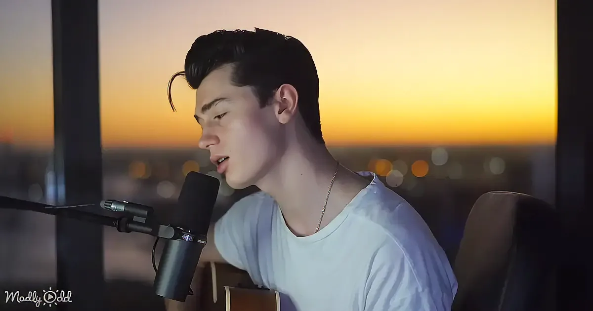 A person sings into a microphone, holding a guitar, with a sunset cityscape in the background.