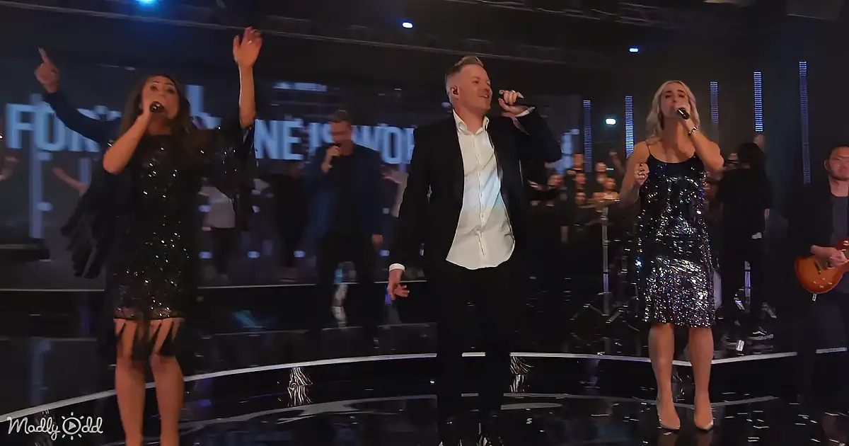 A group of performers in black outfits sing on stage, with one raising their hand and another holding a microphone.