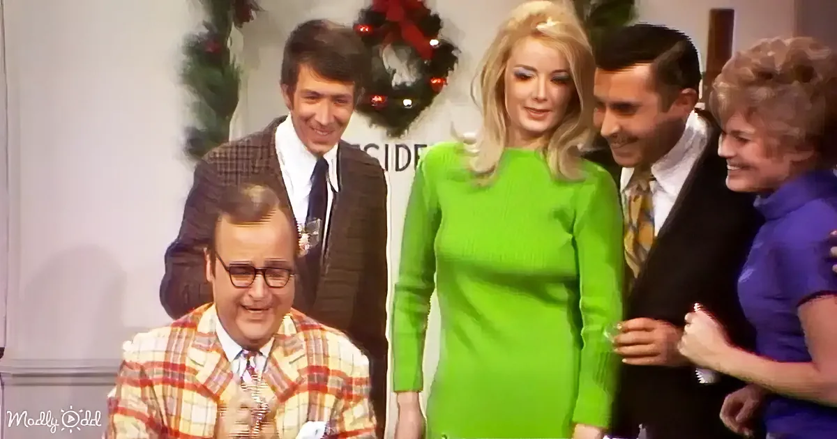 A woman in a bright green dress stands smiling, surrounded by people in formal attire, with a festive wreath in the...