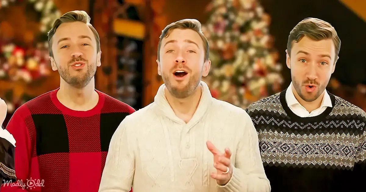 A man in a white sweater gestures while singing, flanked by two others in festive sweaters, with a Christmas tree in the...
