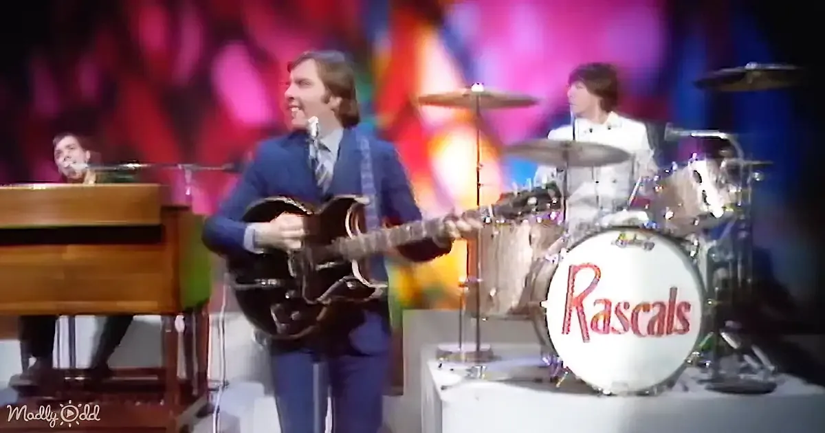 A man in a blue suit plays guitar on stage with a colorful background and a drummer behind him.