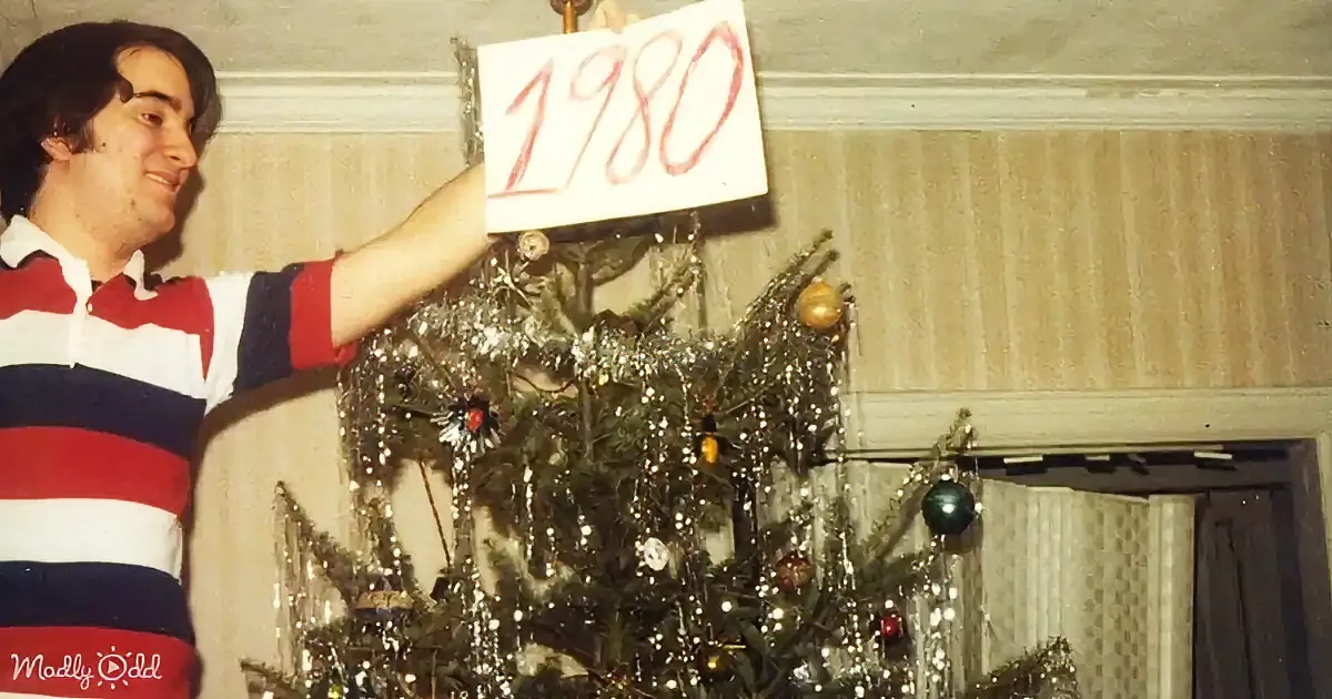 A man in a striped shirt holds up a sign saying "1980" in front of a decorated Christmas tree.