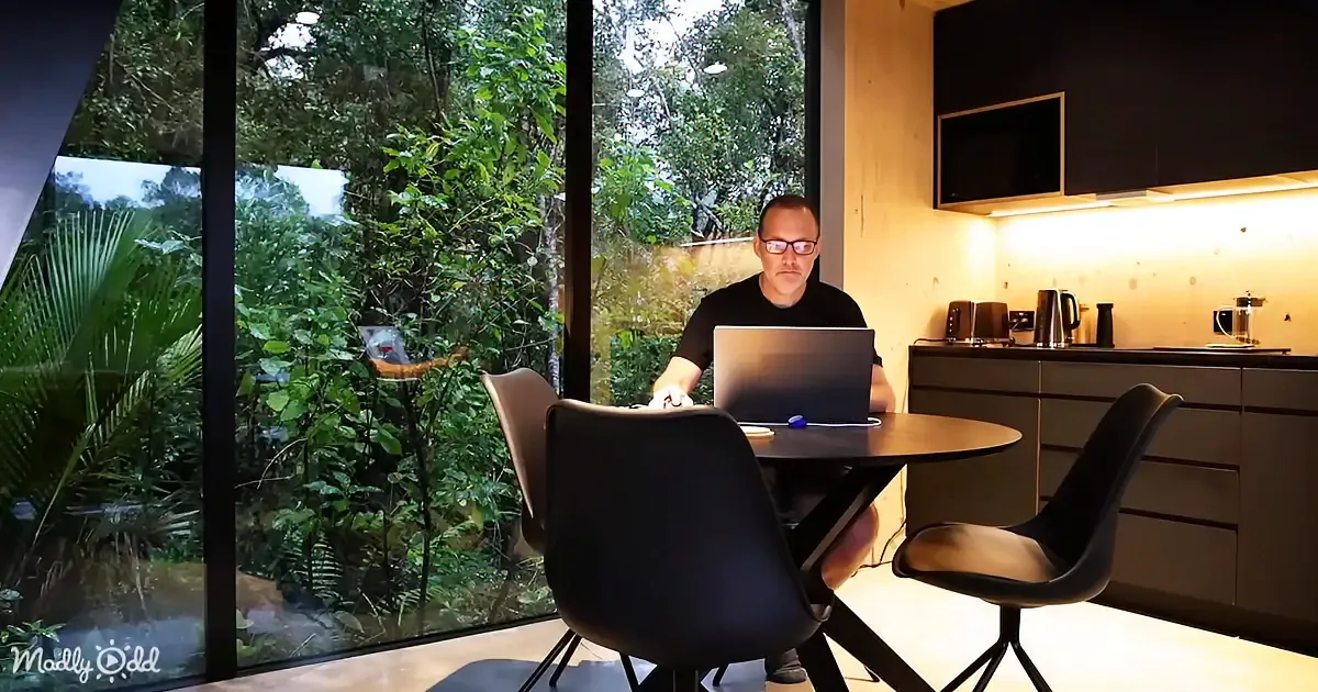 A man sits at a table with a laptop, surrounded by a modern kitchen and large windows showing lush greenery outside.