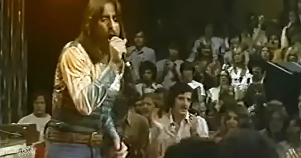 A man in a striped shirt sings into a microphone on stage, surrounded by an audience.