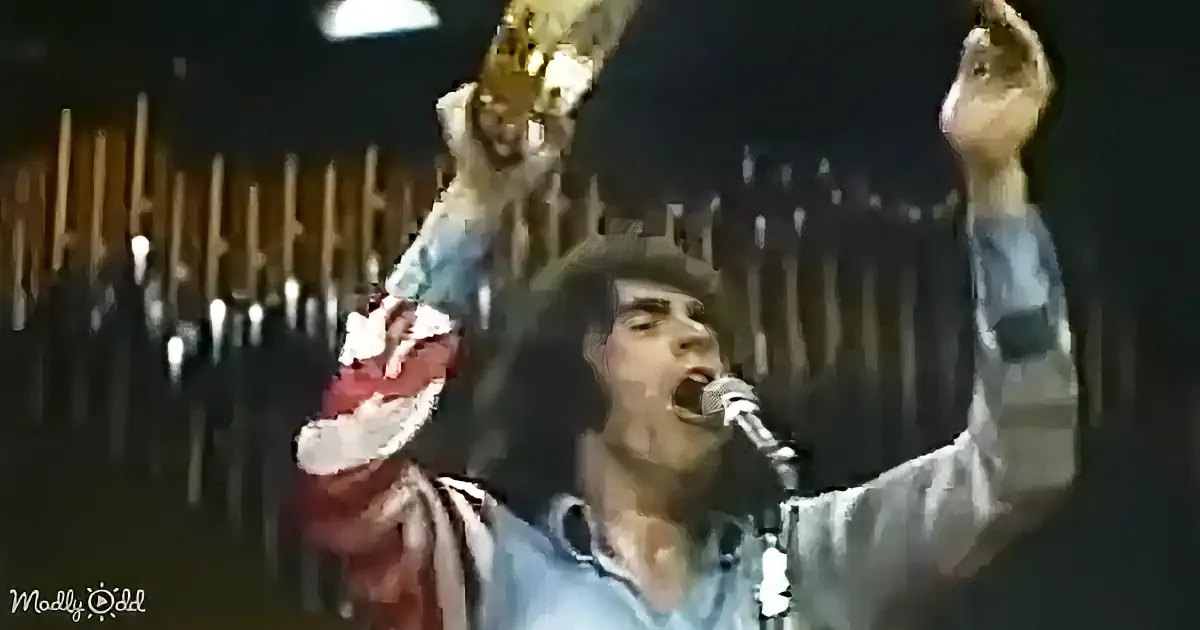 A person in a blue shirt and hat is singing into a microphone with arms raised, holding a red object.