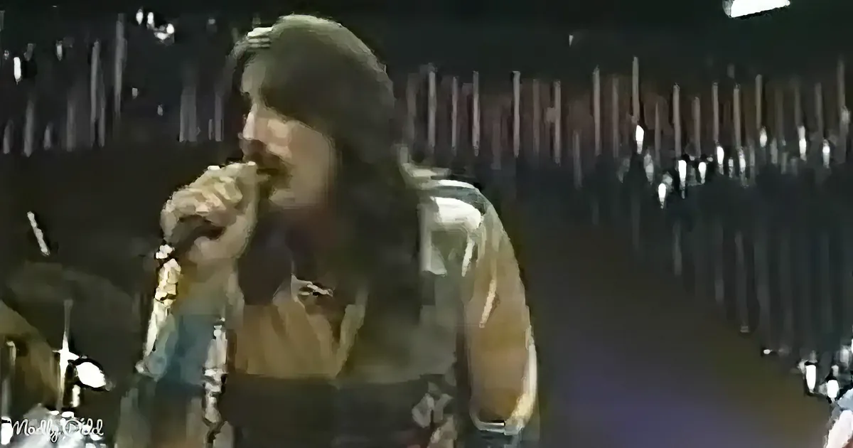 A person with long hair and a leather jacket sings into a microphone on stage.