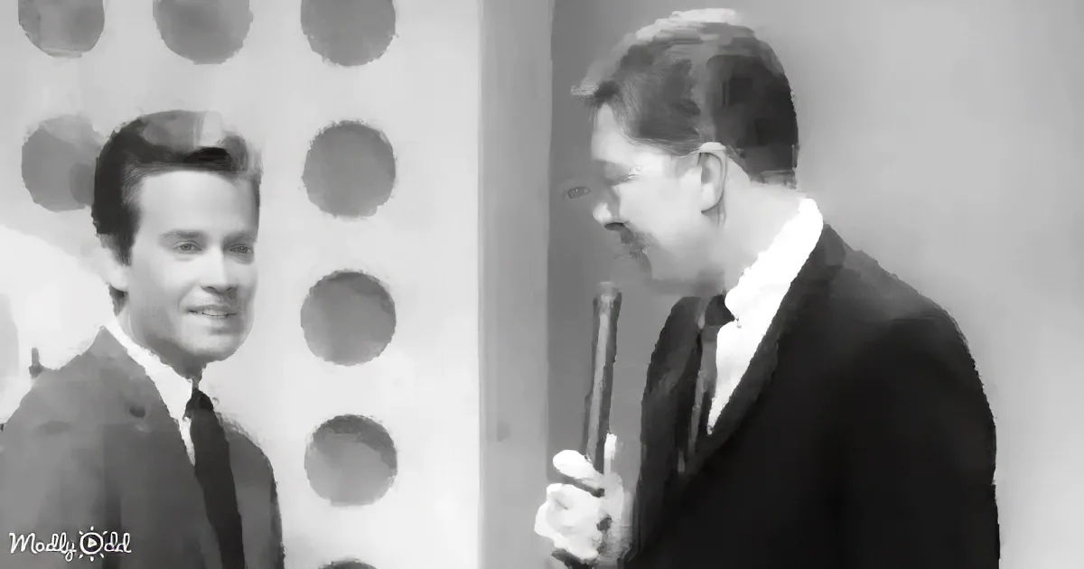 A man in a suit holds a microphone, smiling, with another man in a suit standing nearby.