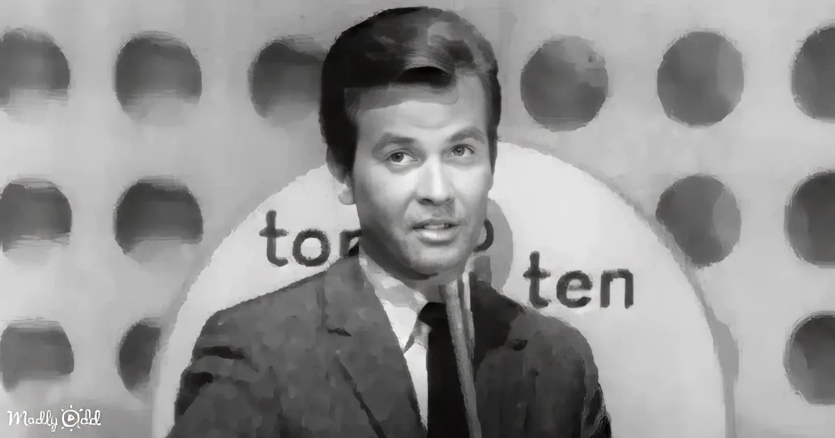 A man in a suit stands in front of a "Top Ten" backdrop, likely hosting a show.