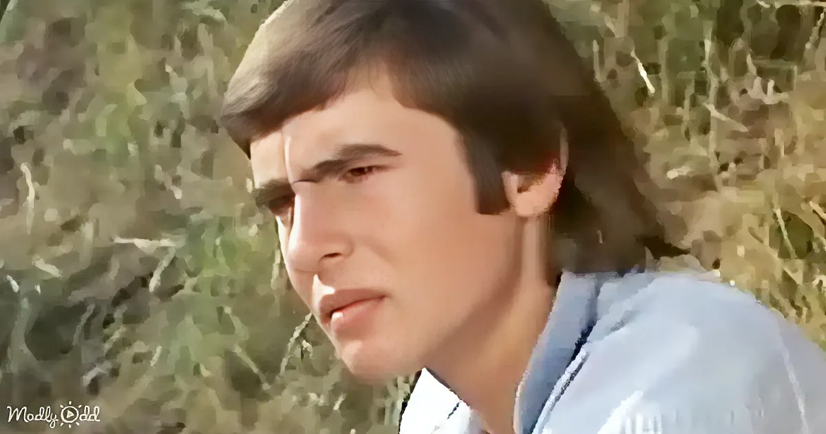 A man with short brown hair, wearing a light blue shirt, is seen in a close-up shot against a blurred natural background.