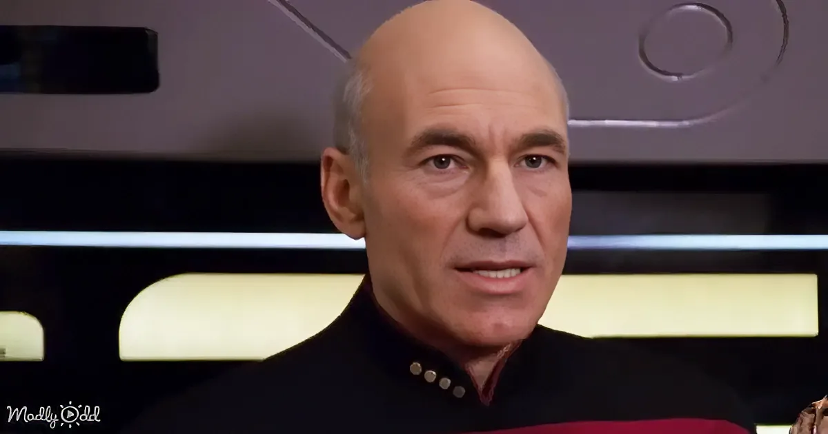 A bald man in a black uniform with red stripes is speaking in a futuristic setting.