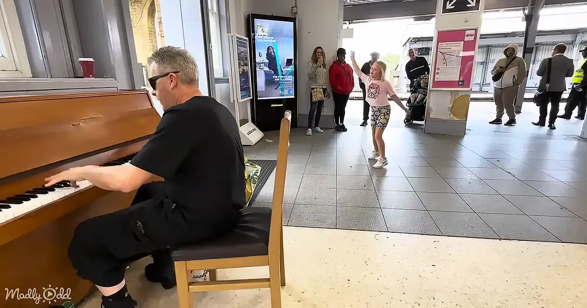 A man plays piano while a girl sings in a public space, surrounded by onlookers.