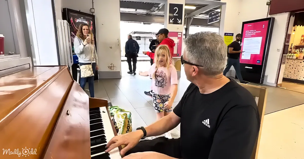 A man plays piano as a young girl watches, with people in the background at a public space.