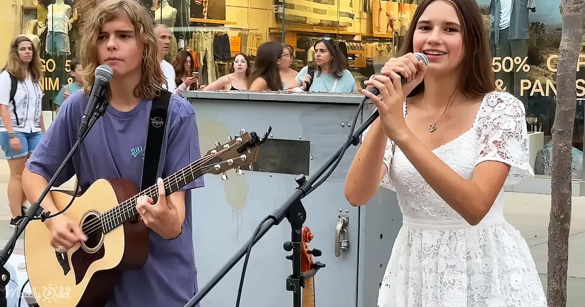 A young man plays guitar while a young woman sings into a microphone in a public setting.