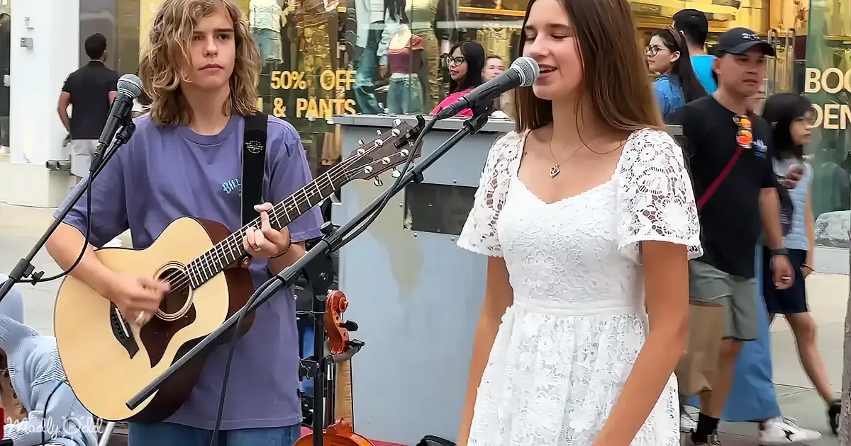 A young woman in a white dress sings into a microphone while a boy plays guitar in front of a store window.