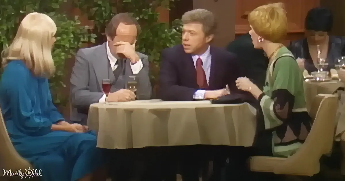 A man in a suit sits at a table with a woman in a blue dress, surrounded by other people in a restaurant setting.