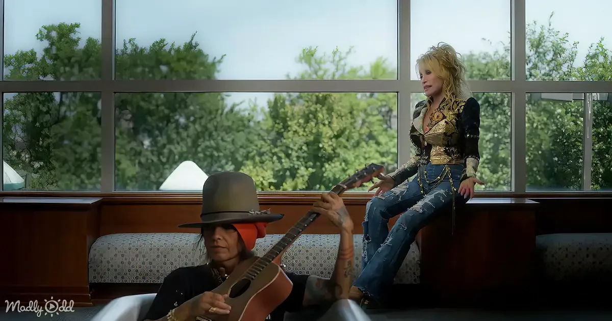 A woman in a cowboy hat plays guitar while another in a gold vest sits on a bench, both in front of large windows.