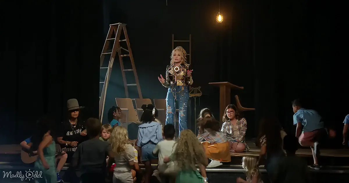 A woman in a denim outfit sings into a microphone on stage, surrounded by children and adults.