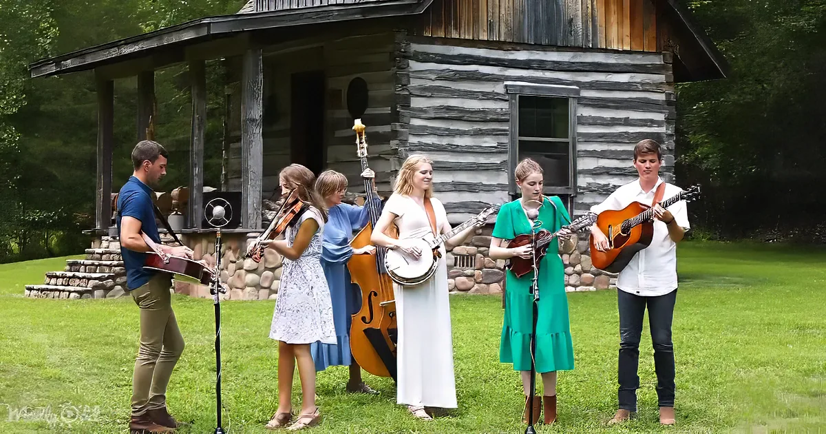 A group of musicians plays traditional instruments in front of a rustic log cabin on a grassy lawn.