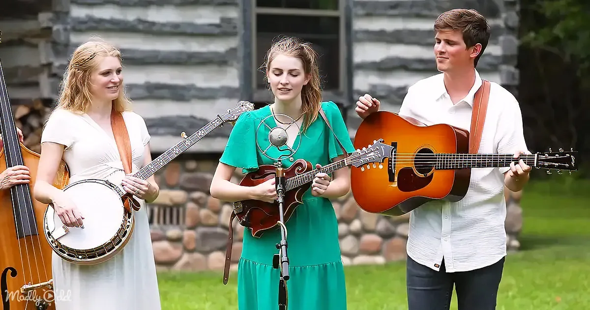 A woman in a green dress plays a mandolin while a man in a white shirt plays a guitar, both in front of a stone building.