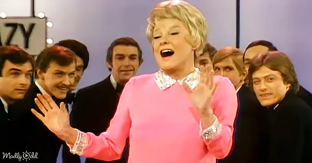 A woman in a pink sweater and floral collar is clapping, surrounded by men in suits, with a sign partially visible in the...
