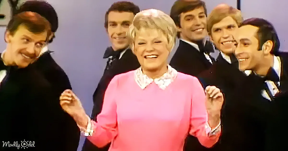 A woman in a pink sweater smiles with a group of men in dark suits behind her.