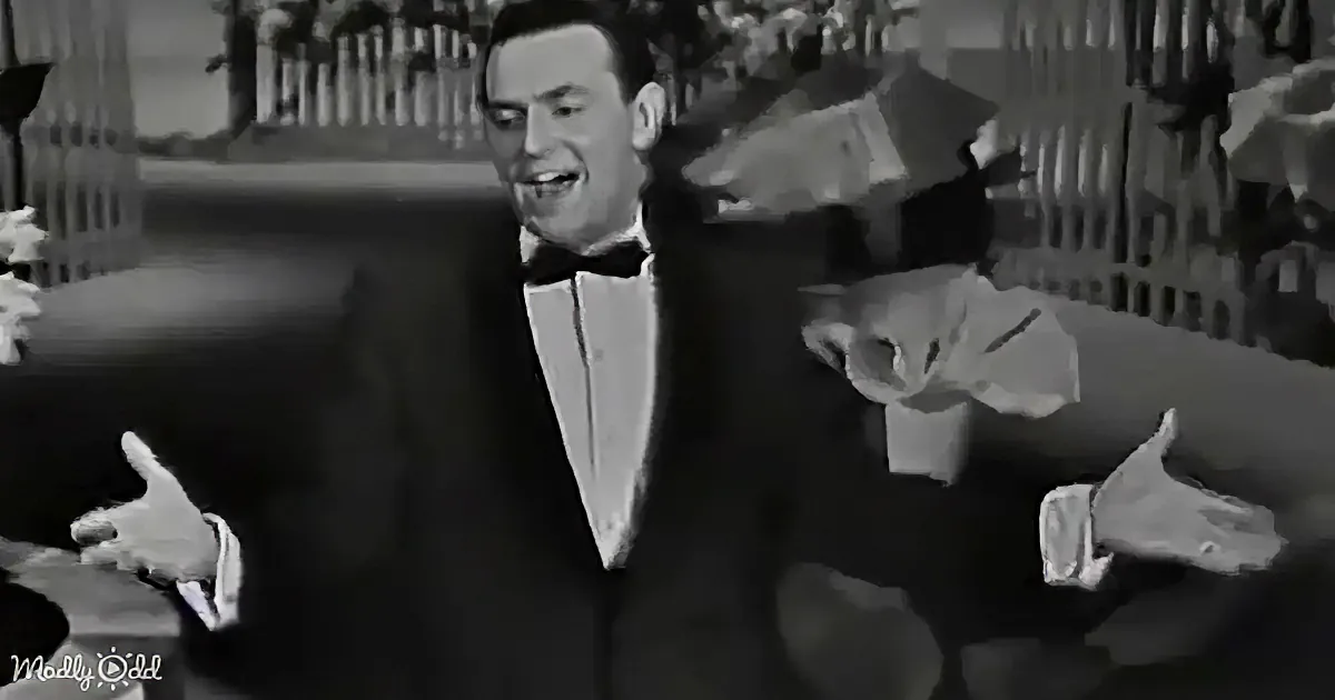A man in a tuxedo and bow tie is smiling, with a blurred background and a hand gesture.