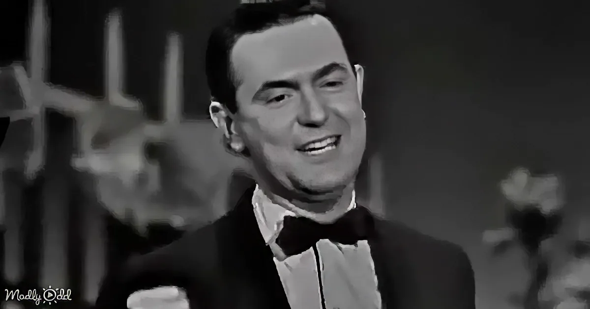 A man in a tuxedo smiles, holding a drink, with a blurred background.