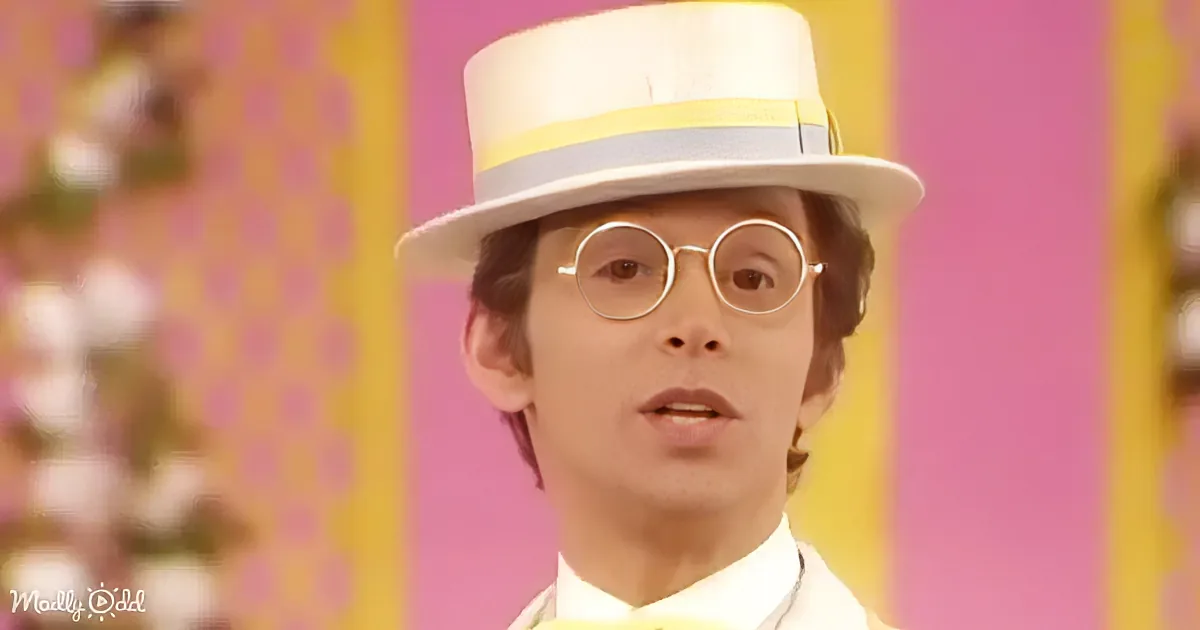 A person wearing a white hat with yellow and gray stripes and round glasses stands in front of a pink and yellow background.