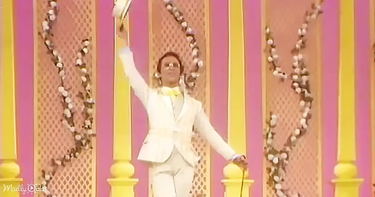 A person in a white suit stands on a stage with arms raised, surrounded by pink and yellow vertical panels.
